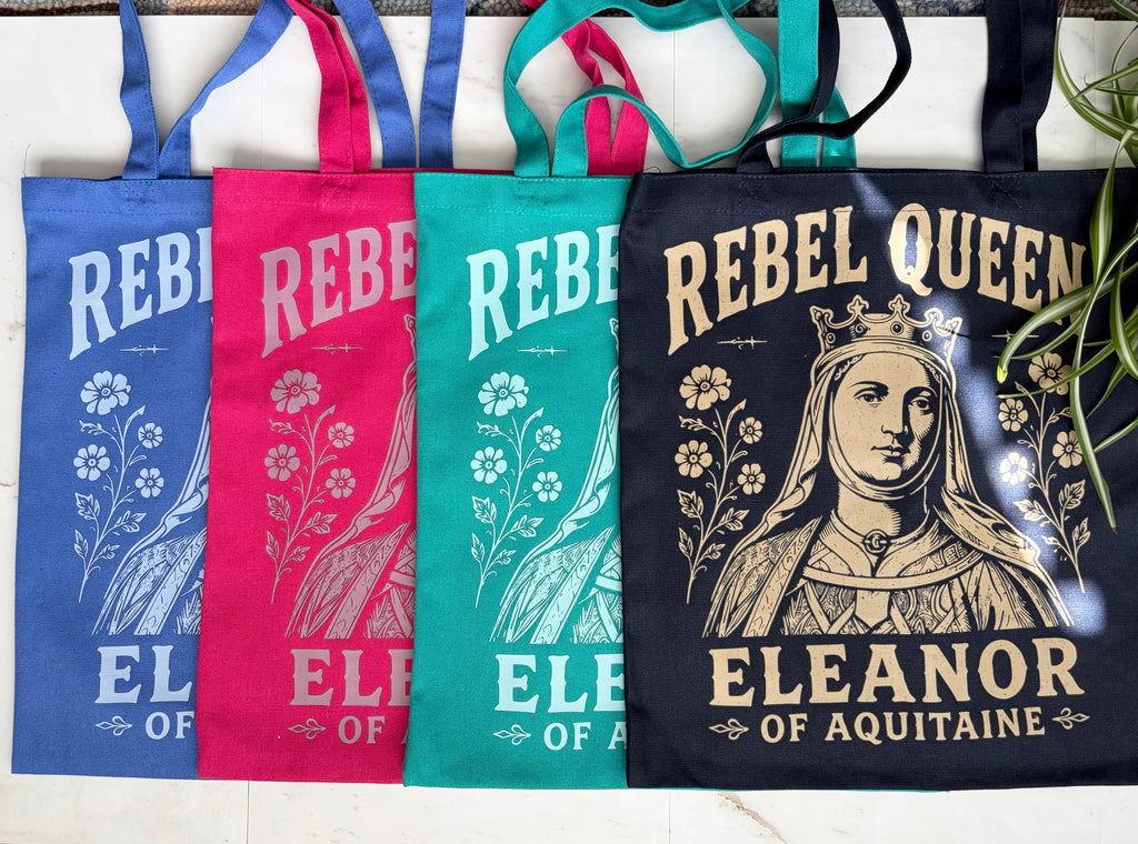 Four fabric tote bags in royal blue, berry pink, teal, and navy, arranged side by side on a white surface. Each bag features a vintage-style graphic with floral illustrations and the text 'REBEL QUEEN' and 'ELEANOR OF AQUITAINA.