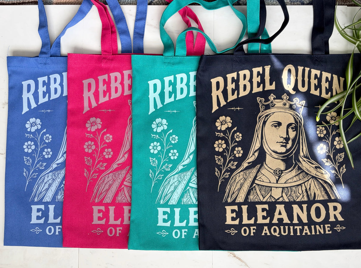 Four fabric tote bags in royal blue, berry pink, teal, and navy, arranged side by side on a white surface. Each bag features a vintage-style graphic with floral illustrations and the text 'REBEL QUEEN' and 'ELEANOR OF AQUITAINA.