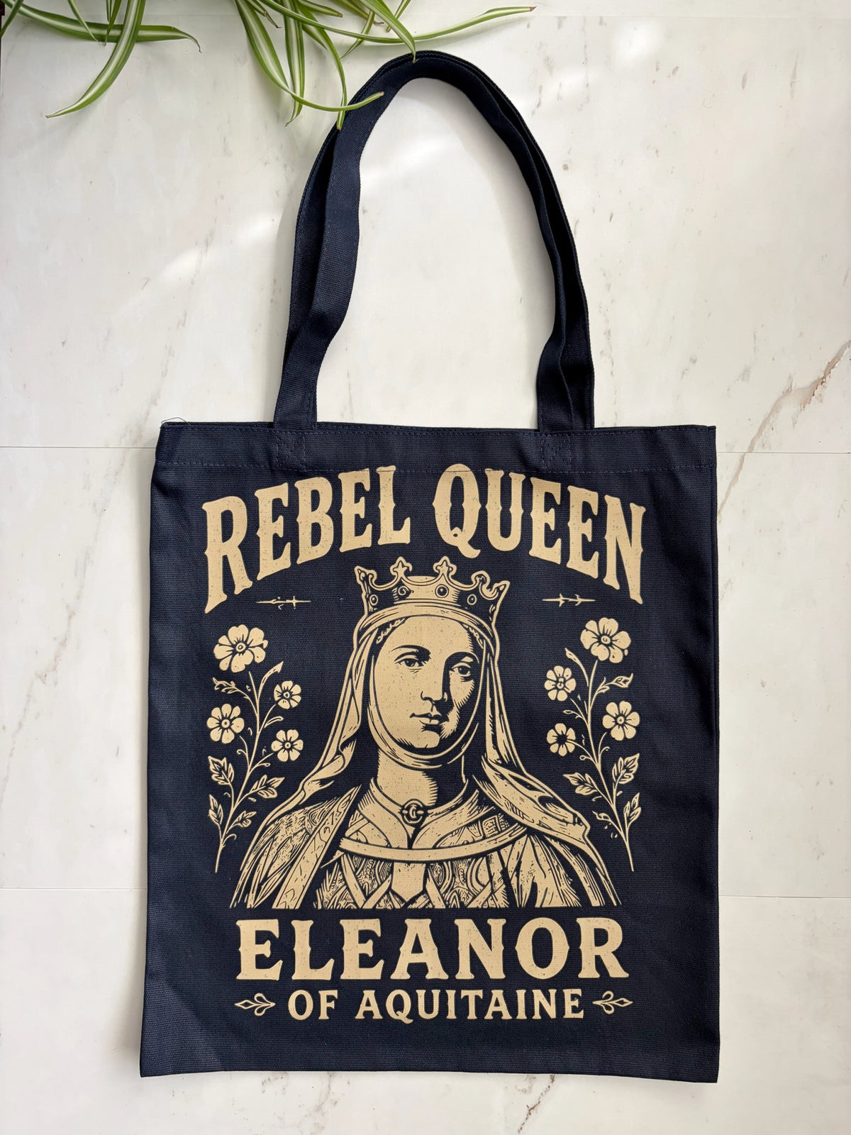A navy blue canvas tote bag with a vintage-style cream graphic of Queen Eleanor of Aquitaine and the text 'REBEL QUEEN' and 'ELEANOR OF AQUITAINE', displayed on a light marble surface with a green plant leaf in the corner.