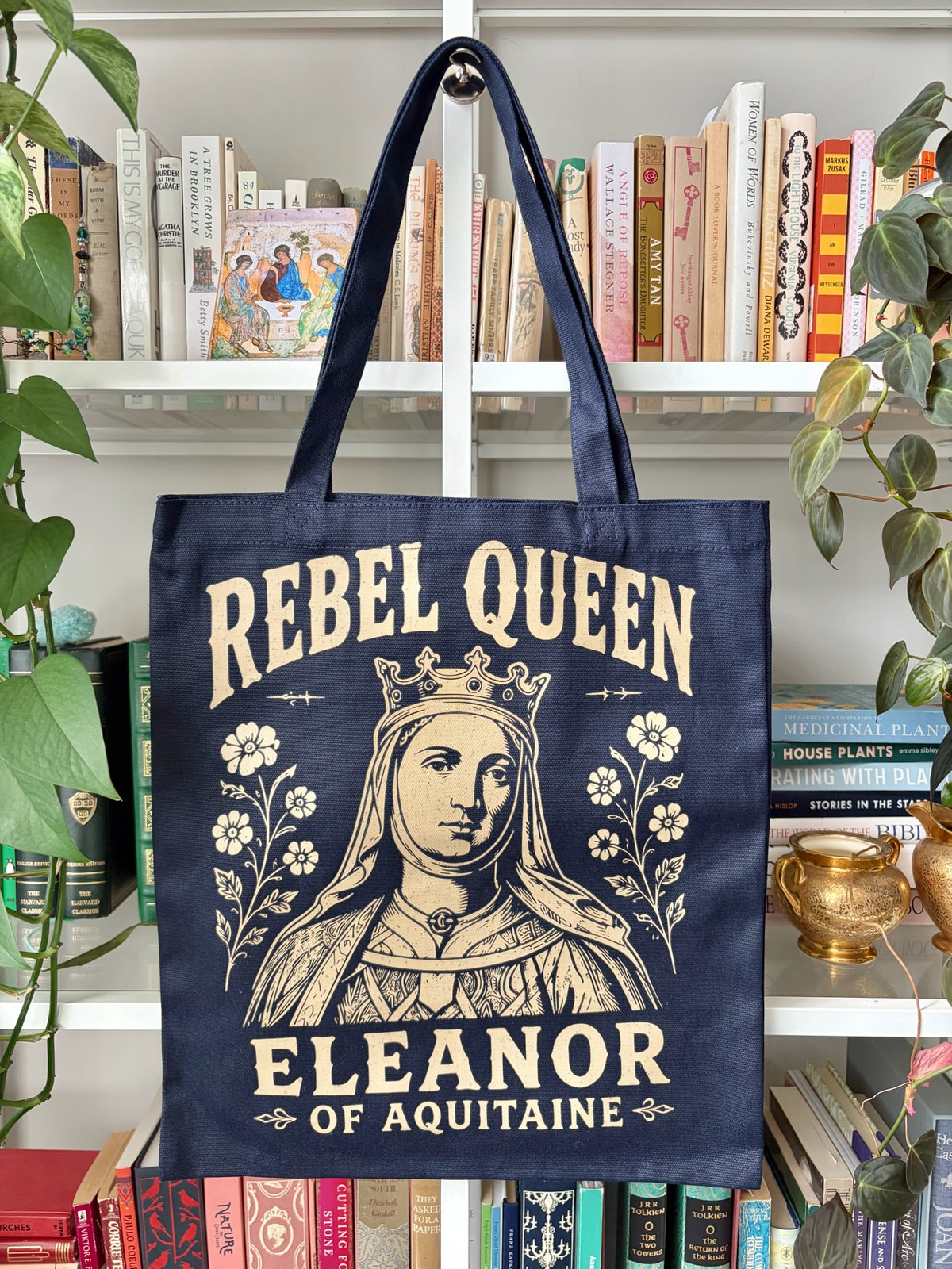A navy blue canvas tote bag with a cream-colored graphic of Queen Eleanor of Aquitaine and the text 'REBEL QUEEN' and 'ELEANOR OF AQUITAINA', hanging on a white bookshelf surrounded by books and green plants.