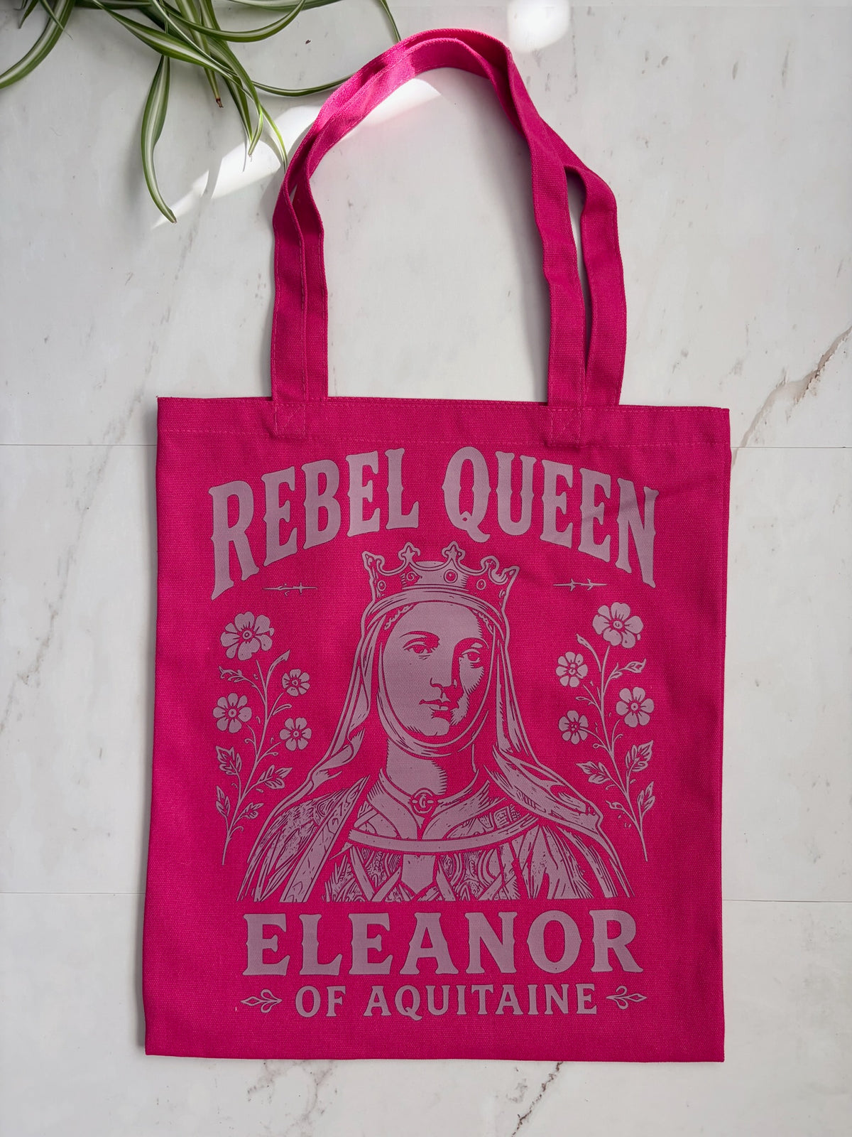 A bright pink berry canvas tote bag with a light pink graphic of Queen Eleanor of Aquitaine and the text 'REBEL QUEEN' and 'ELEANOR OF AQUITAINE', photographed on a light marble surface with a plant leaf in the corner.