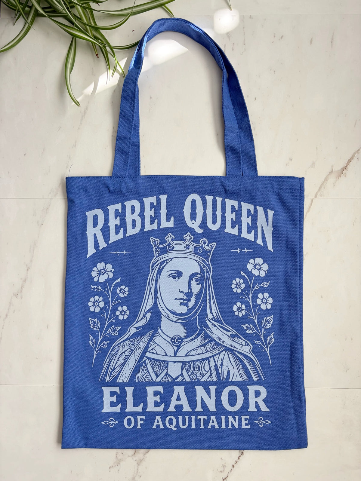 A royal blue canvas tote bag with a white graphic print of Queen Eleanor of Aquitaine and the text 'REBEL QUEEN' and 'ELEANOR OF AQUITAINA', displayed on a light marble surface with a green plant leaf in the corner.