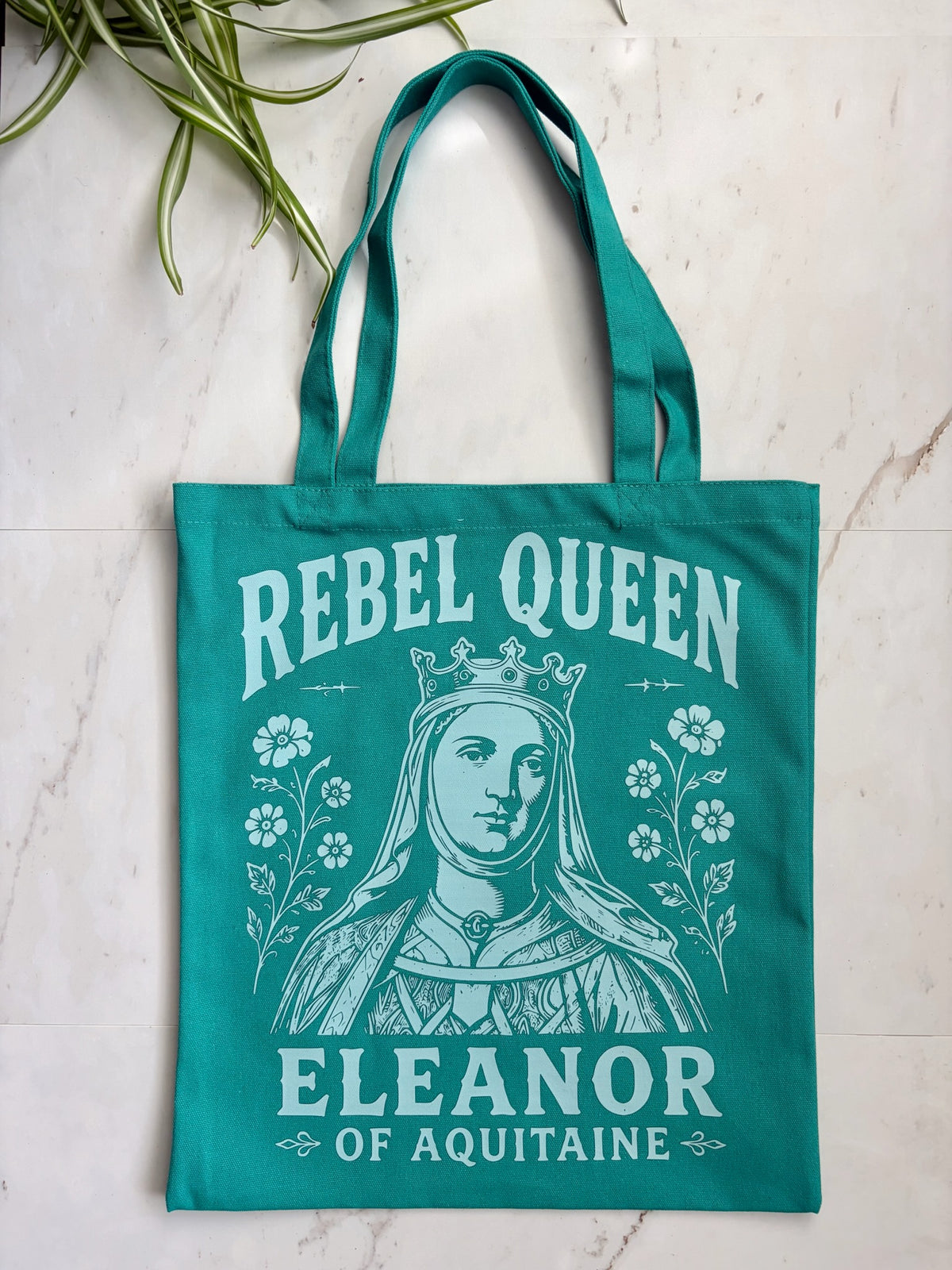 A teal canvas tote bag with a white graphic print of a crowned queen and the text 'REBEL QUEEN ELEANOR OF AQUITAINE', photographed flat on a light marble surface with a plant leaf in the corner.